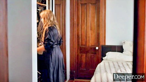 Blonde woman in silky black dress opens closet in bedroom, teasing curvy figure from behind