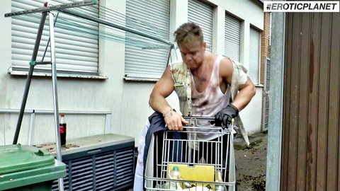 Sweaty muscular blonde hunk in tank top pushes shopping cart in outdoor alley
