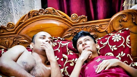 Two South Asian men lying thoughtfully on ornate wooden bed with red pillows and curtains