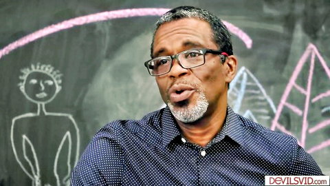 Mature black teacher with glasses speaking passionately in front of chalkboard before taboo student sex.