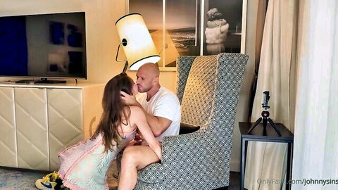 Skinny long-haired brunette in short dress passionately kisses muscular bald man in white tee on hotel armchair POV