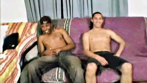 Shirtless white long-haired guy and black BBC dude smiling on couch in interracial gay first-time video
