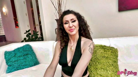 Curly haired brunette MILF smiles seductively with deep cleavage in black halter top on couch