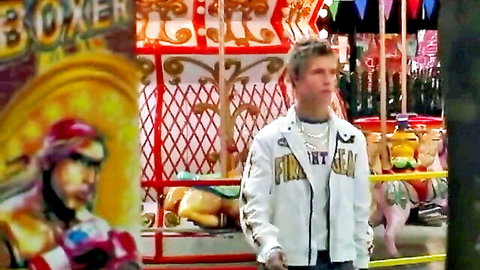 Young blonde guy in white FUN GAME jacket stands at vibrant carnival carousel with boxer clown sign