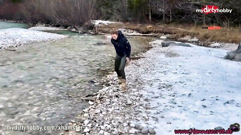 Blonde woman squats in shallow rocky river outdoors with snowy banks and woods