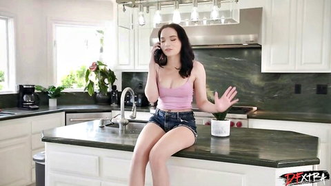 Petite brunette teen in tight pink tank top and denim shorts sitting seductively on kitchen counter on phone