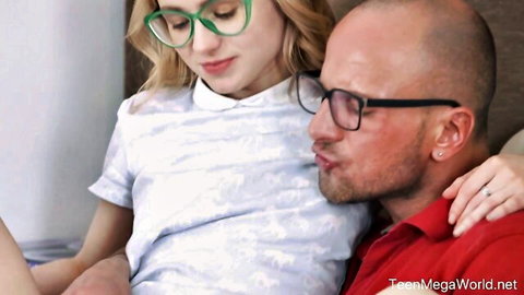 Blonde teen with glasses sits intimately on bald man's lap in white blouse