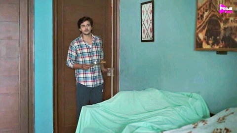 Handsome Indian man in plaid shirt standing by bedroom door with green bed sheets