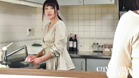 Cute Japanese babe in beige dress washing dishes smiling at another woman from behind in kitchen