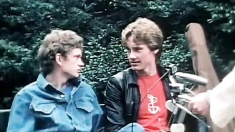 Two young men in denim and leather jackets chatting on a bench beside motorcycle in vintage outdoor scene