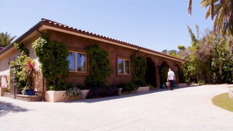 Man in white shirt walking away from luxurious stucco house with suitcase on sunny driveway
