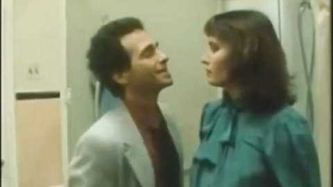 Vintage brunette MILF in teal bow blouse stares intimately at suited curly-haired man in bathroom