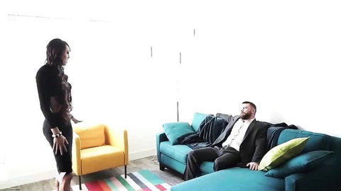 Sexy black woman in tight cutout dress stands dominantly near suited bearded man lounging on sofa