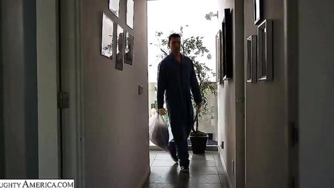 Handyman in blue jumpsuit walks down sunlit apartment hallway carrying white bag