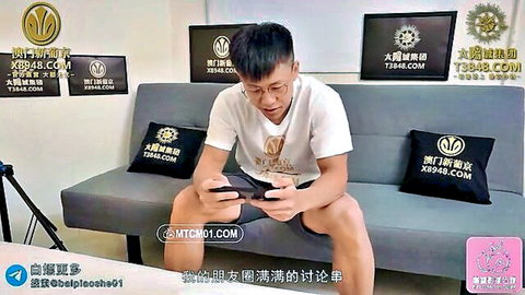 Young Asian man in white shirt and shorts sits on gray couch holding phone in live stream studio