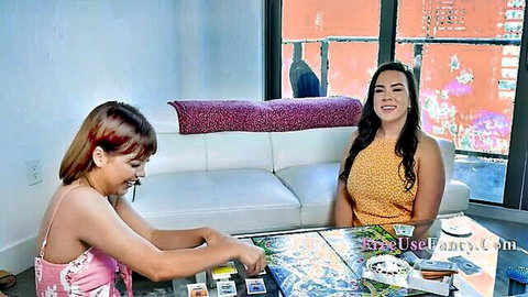 Two smiling Latinas in skimpy tops playing naughty board game on couch