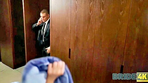 Suited bald groom peeking from wooden door grabbing bride's blue dress on wedding hotel floor