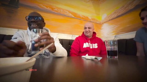 Black man in sunglasses and bald guy in red hoodie smiling with blonde girl at bar table drinking
