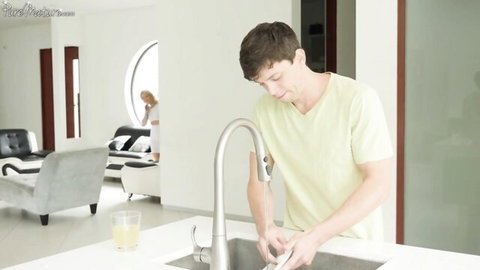 Young man washes dishes in kitchen while blonde woman sits in background living room