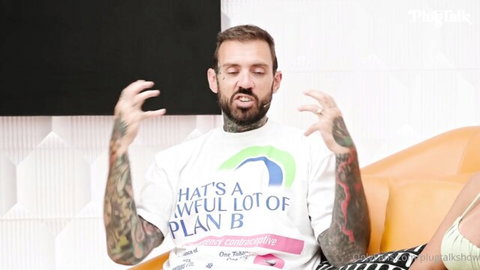 Tattooed man in 'That's a whole lot of Plan B' t-shirt gesturing excitedly on orange couch