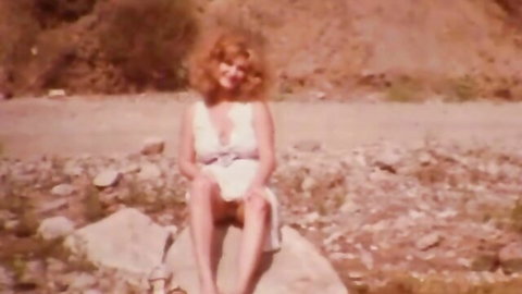Retro redhead in low-cut white dress sits on rock outdoors showing natural cleavage