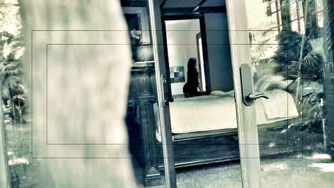 Seductive MILF with dark hair sitting on bed, viewed through doorframe