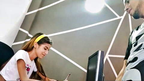 Skinny Latina girl in tight white t-shirt smiles at desk while guy leans over her