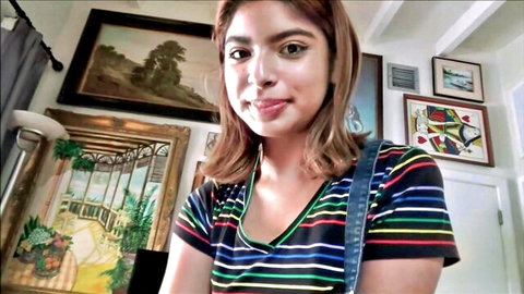 Brunette teen smiling seductively in colorful striped V-neck top in art-filled room