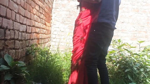 Desi woman in red saree embraced by man in outdoor setting