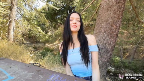 Hot black-haired teen babe in off-shoulder blue top smiling seductively against tree in forest