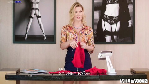Blonde MILF Sydney Paige holds up red thong panties in front of lingerie poster