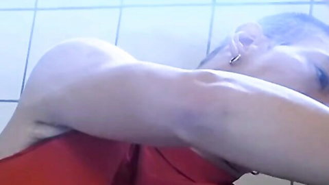 Young muscular gay man with ear piercing stretches arm sensually in red top bathroom