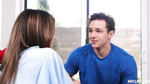 Long-haired brunette MILF in light blue top smiling at handsome guy in blue ribbed shirt by window