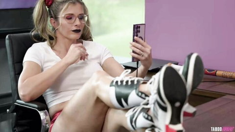 Blonde cheerleader in tight white crop top and red shorts spreads legs wide checking phone on chair