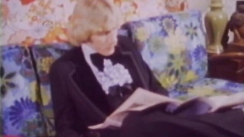 Blonde man in tuxedo reading newspaper on retro floral couch