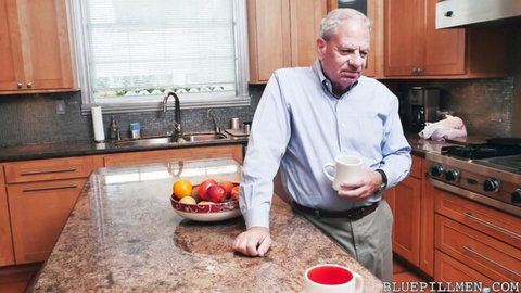 Mature gray-haired man in blue shirt holding coffee mug leaning on kitchen counter