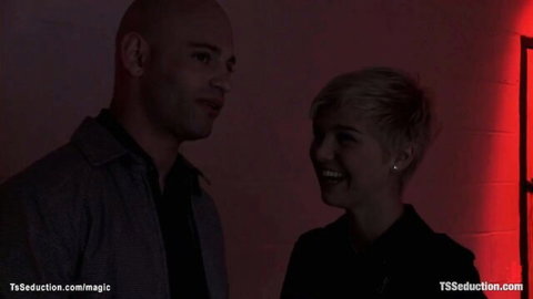 Blonde TS seduces smiling bald man in close intimate chat, teasing threesome vibe