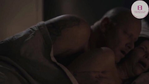 Tattooed bald man spoons woman passionately in dim bedroom under sheets
