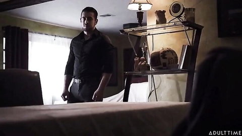Handsome man in black shirt stands in dimly lit bedroom with bed and shelves, ready for steamy action.