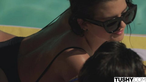 Tanned skinny brunette lies topless in black thong on pool lounger as young stud kisses her back