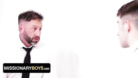Older suited daddy stares intensely at young twink in white shirt and tie in gay Missionary Boys scene