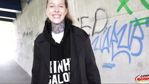 Young blonde with neck tattoo smiles in graffiti tunnel wearing black hoodie