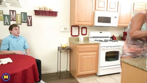 Tanned MILF in sheer orange apron exposing thick ass and pussy to young guy at kitchen table