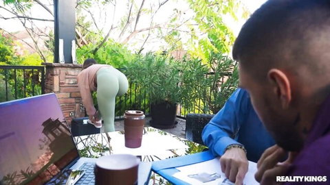 Curvy woman in tight green yoga pants bends over outdoor table showing round ass to tattooed man on laptop