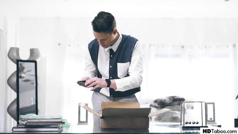 Suited brunette man checking phone while unboxing package on glass desk in modern office