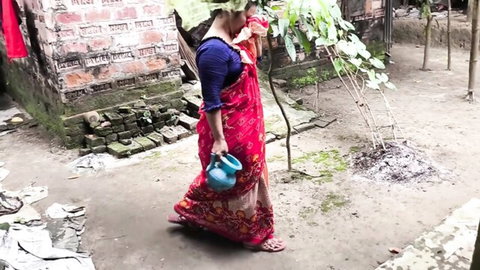 Desi Indian stepmom in red sari holding blue pitcher, walking in rural outdoor setting