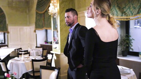 Seductive blonde in tight black dress puckers lips looking back at suited man in elegant dining room