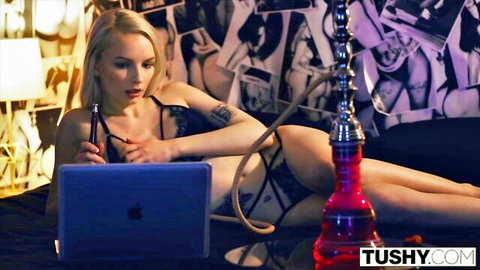 Blonde babe in sheer black lingerie seductively smokes hookah while lounging with laptop