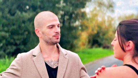 Tattooed bald man seduces busty brunette babe outdoors gripping her shoulder intimately