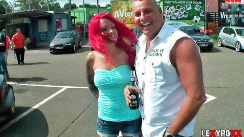 Busty tattooed redhead in tight turquoise tube top and denim shorts poses smiling with muscular bald guy holding beer in parking lot